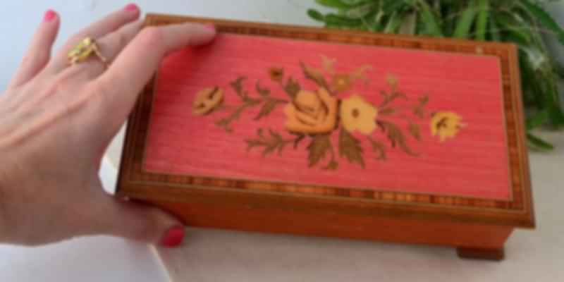 A person's hand opening the lid of a red, ornately painted wooden music box.