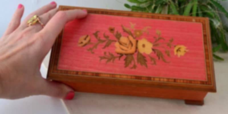A person's hand opening the lid of a red, ornately painted wooden music box.